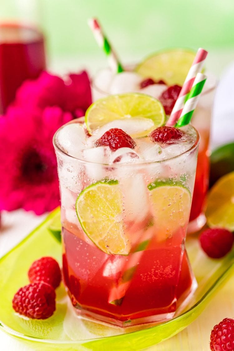 A glass filled with a raspberry lime rickey on a green serving tray with raspberries around it. 