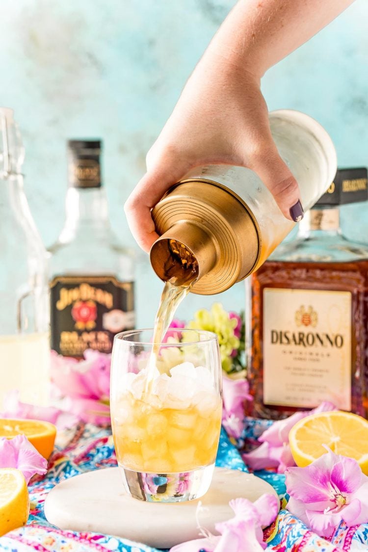 A cocktail shaker pouring a drink into a rocks glass filled with ice.