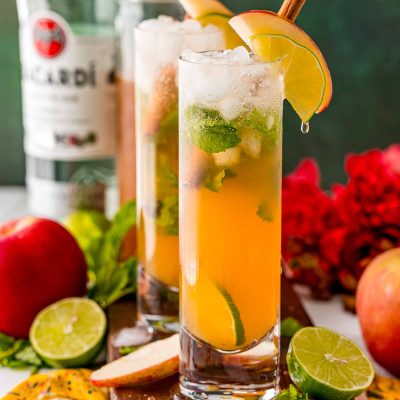 Close up photo of a couple of apple mojitos on a wooden serving tray surrounded by limes and apples.