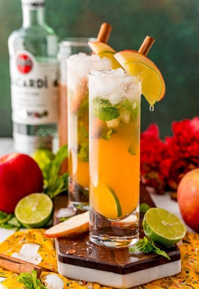 Close up photo of a couple of apple mojitos on a wooden serving tray surrounded by limes and apples.