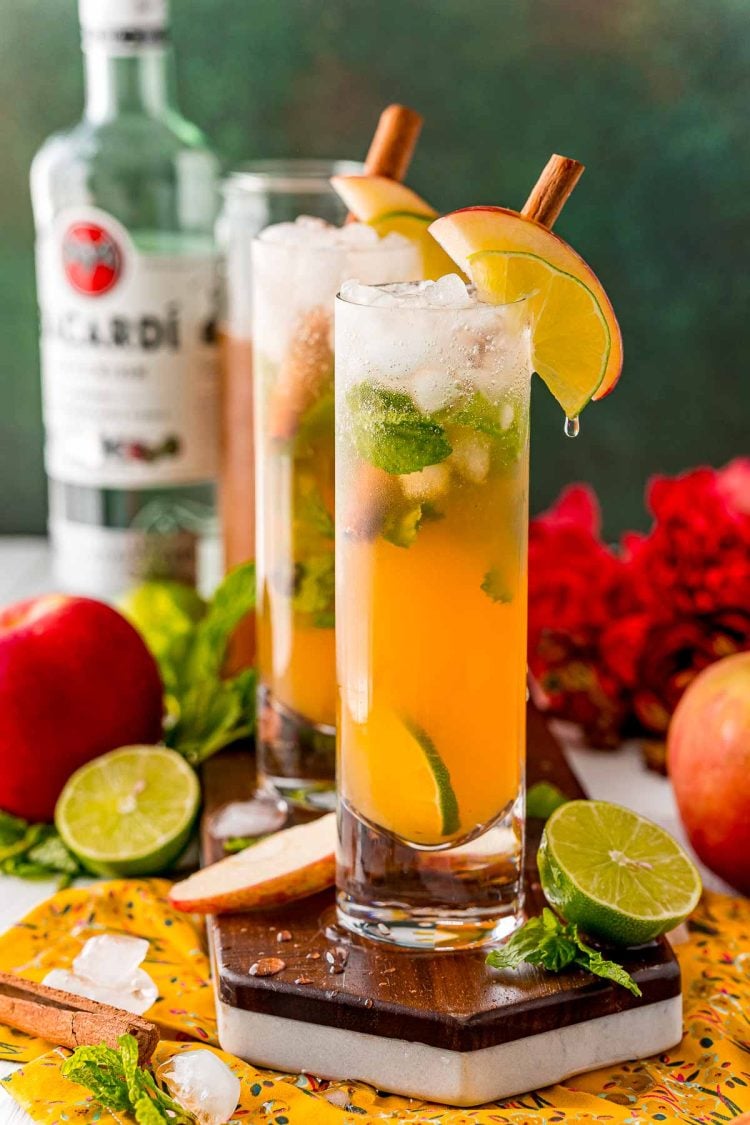 Close up photo of a couple of apple mojitos on a wooden serving tray surrounded by limes and apples.