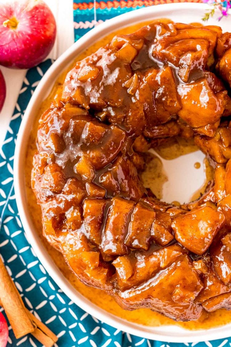 Overhead photo of apple monkey bread on a white plate on a blue napkin with cinnamon sticks and apples in the background.