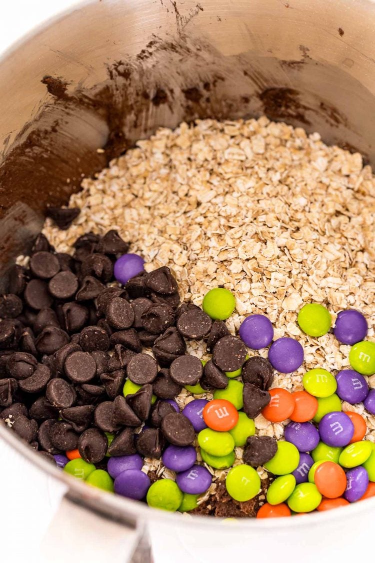 Oatmeal, chocolate chips, and M&Ms being added to a mixing bowl.