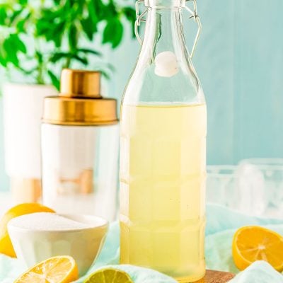 Bottle of homemade sour mix with lemons and limes around it and a cocktail shaker in the background.