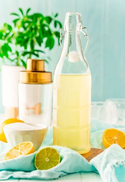 Bottle of homemade sour mix with lemons and limes around it and a cocktail shaker in the background.