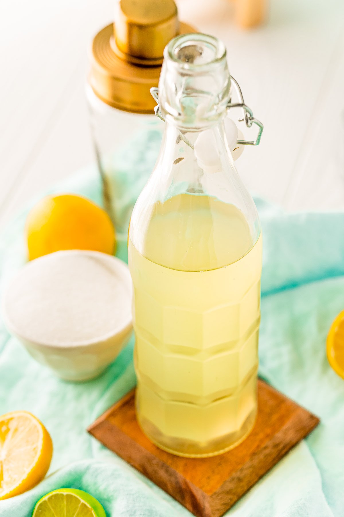 Bottle of homemade sour mix with lemons and limes around it and a cocktail shaker in the background.