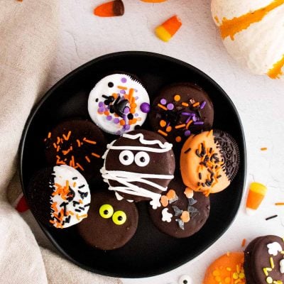 Overhead photo of halloween chocolate covered oreos on a black plate surrounded by pumpkins and candy corn.