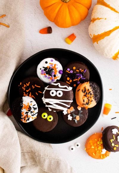 Overhead photo of halloween chocolate covered oreos on a black plate surrounded by pumpkins and candy corn.