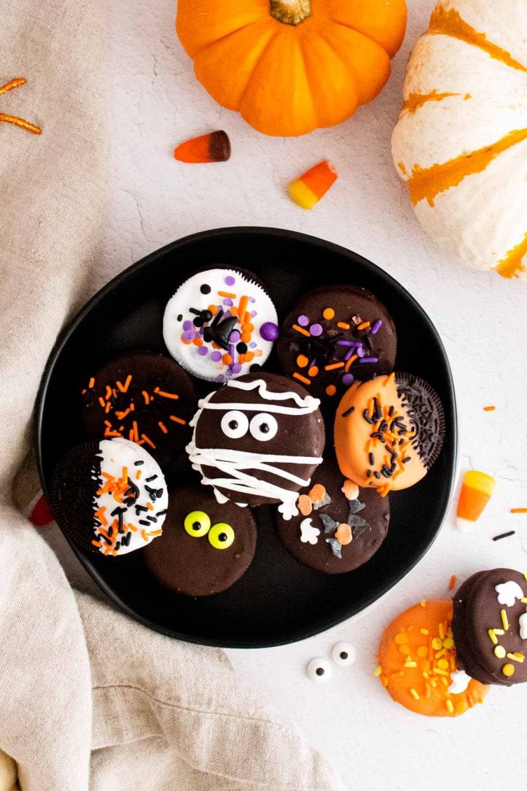 Overhead photo of halloween chocolate covered oreos on a black plate surrounded by pumpkins and candy corn.