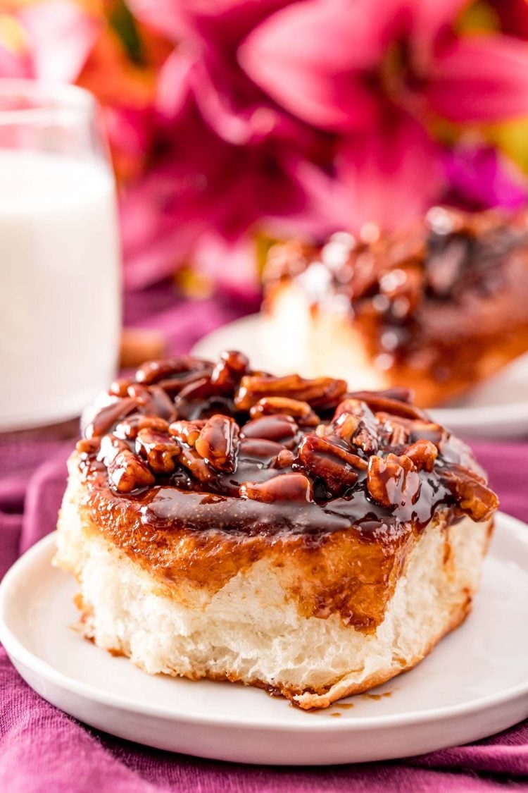 Cinnamon pecan sticky bun on a small white plate with pink flowers and a glass of milk in the background.