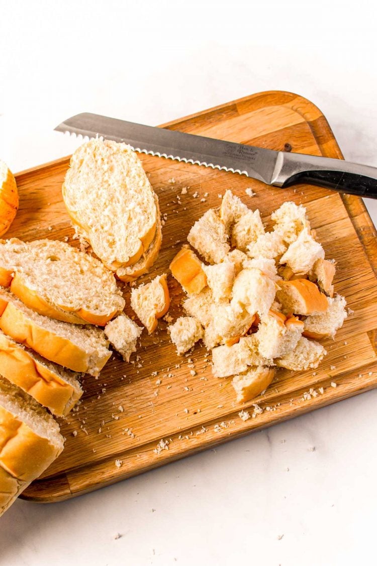 French bread being cut into cubes for stuffing on a wooden cutting board.