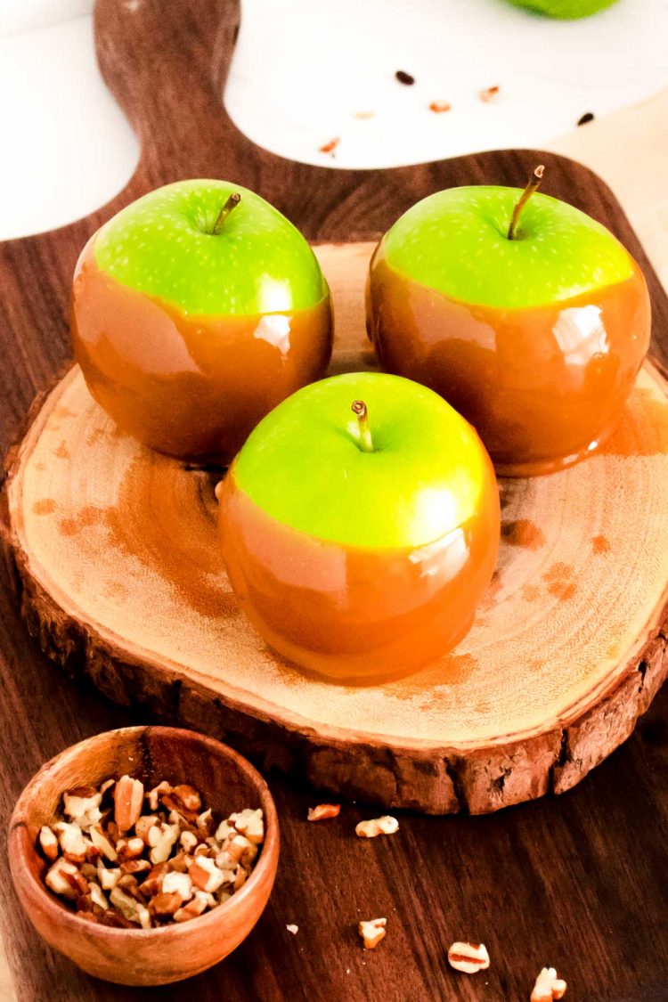 Three caramel dipped apples on a wooden cutting board surrounded by nuts.