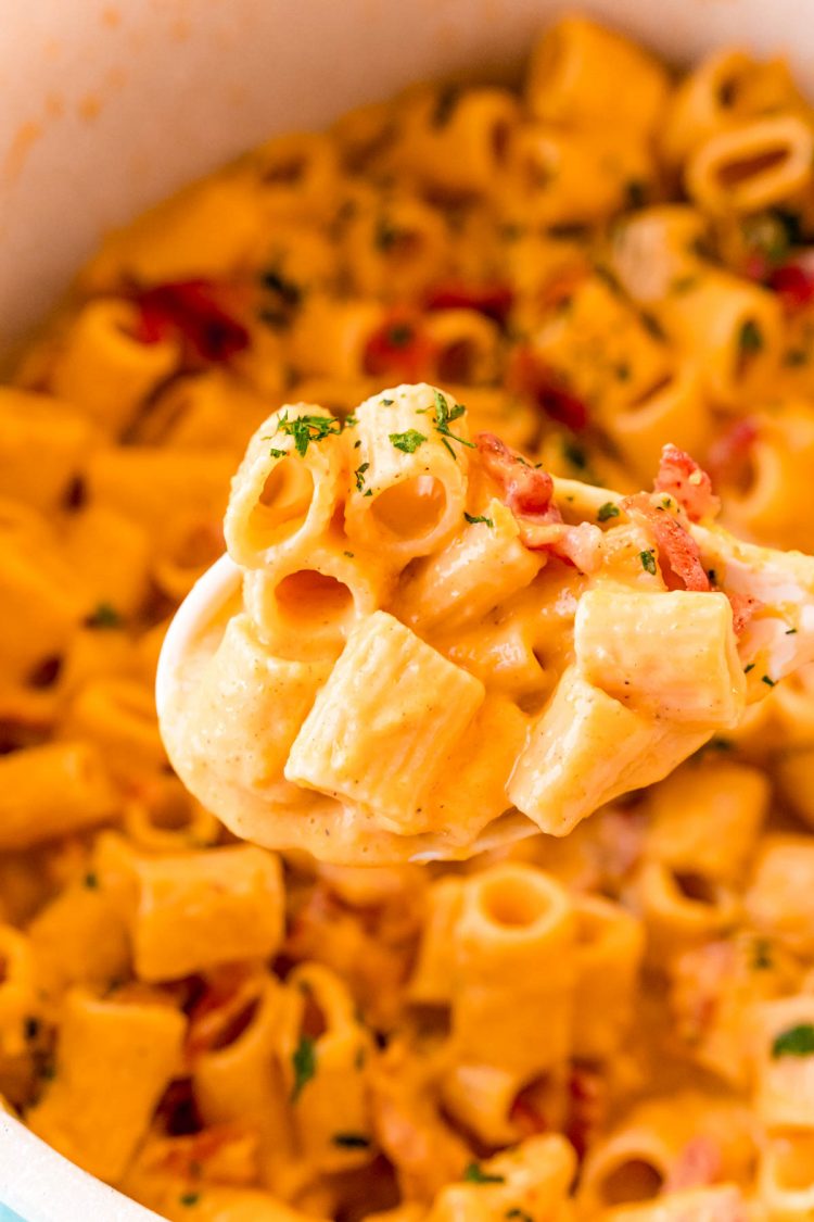 A white serving spoon lifting a scoop of mac and cheese with bacon out of a pot.