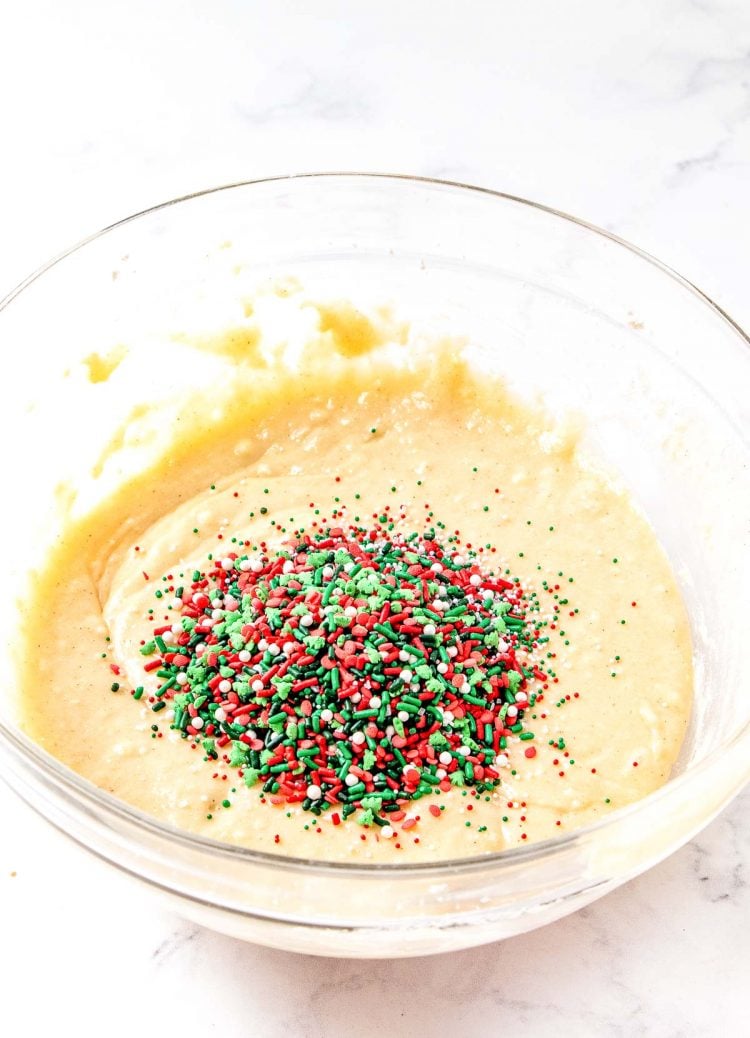Christmas sprinkles being added to a glass mixing bowl filled with batter for quick bread.