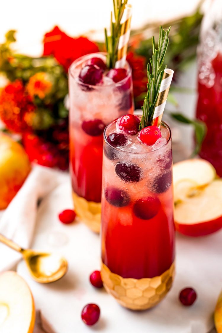 Close up photo of two red cranberry cider drinks on a white table with apples and cranberries scattered around.