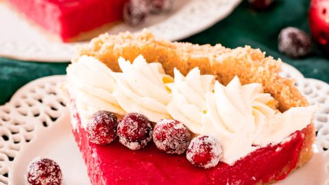 A slice of cranberry pie on a white plate topped with whipped cream and sugared cranberries.