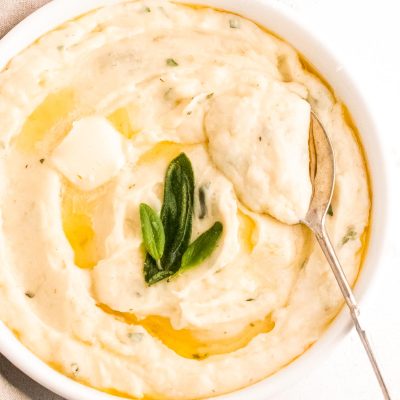 Close up photos of garlic mashed potatoes topped with butter and sage in a white serving dish with a serving spoon in them.