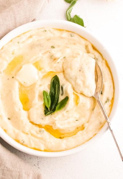 Close up photos of garlic mashed potatoes topped with butter and sage in a white serving dish with a serving spoon in them.