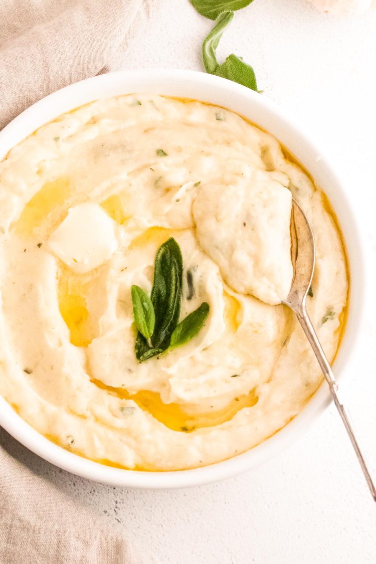 Close up photos of garlic mashed potatoes topped with butter and sage in a white serving dish with a serving spoon in them.