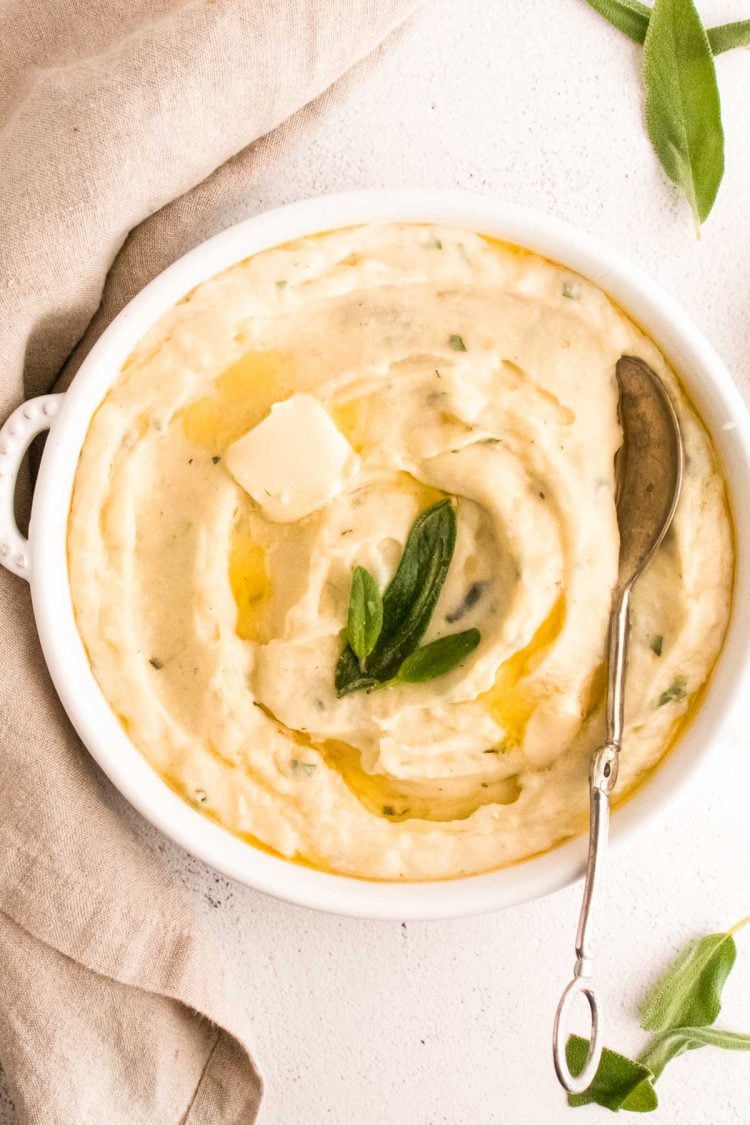 Close up photos of garlic mashed potatoes topped with butter and sage in a white serving dish.