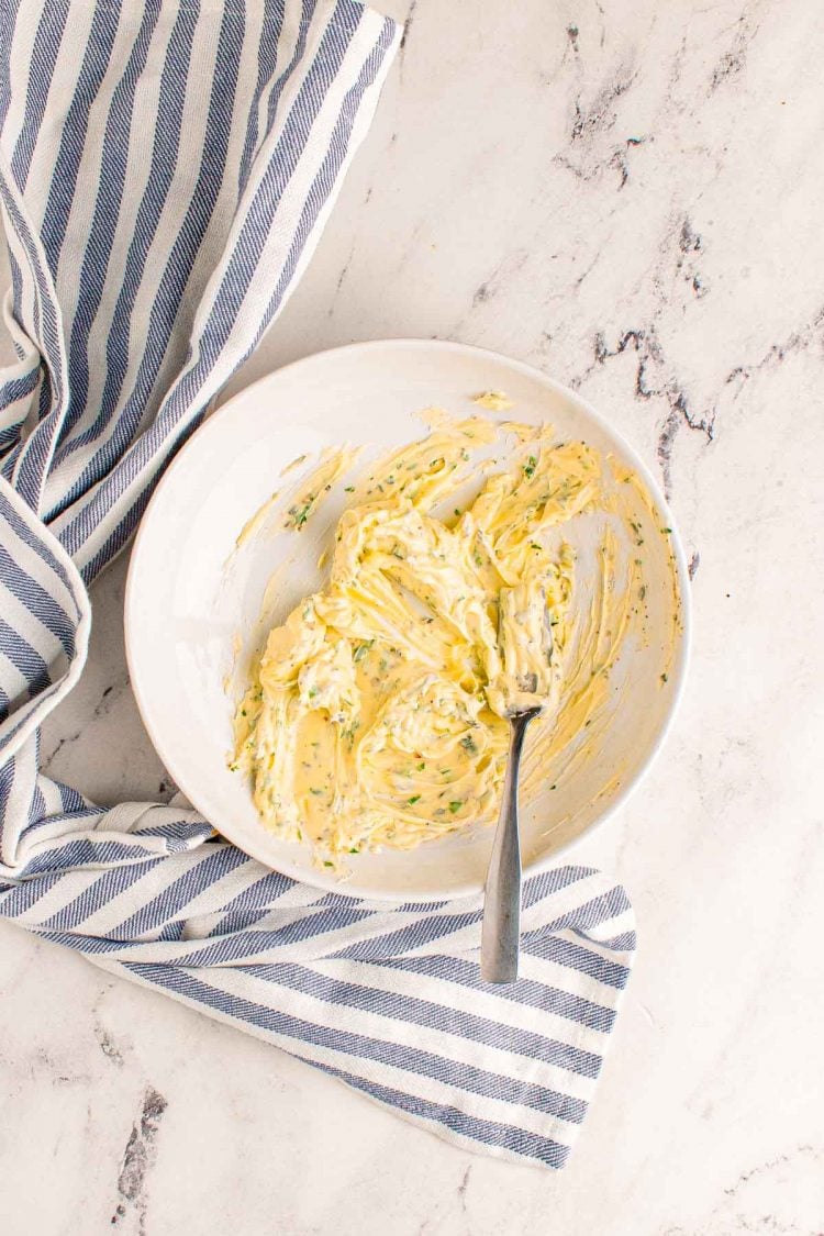 Herb butter being mixed with a fork in a white bowl on a marble surface.