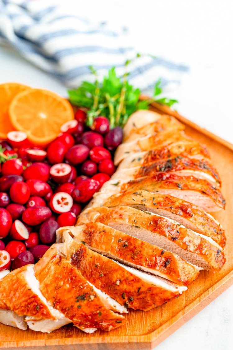 Close up of a sliced turkey breast on a wooden plate with cranberries, orange slices, and herbs for garnish.