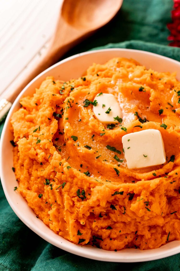 Close up photo of mashed sweet potatoes topped with butter and parsley in a white bowl.