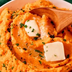 Close up photo of a bowl of mashed sweet potatoes topped with butter with a wooden spoon taking a scoop out of it.