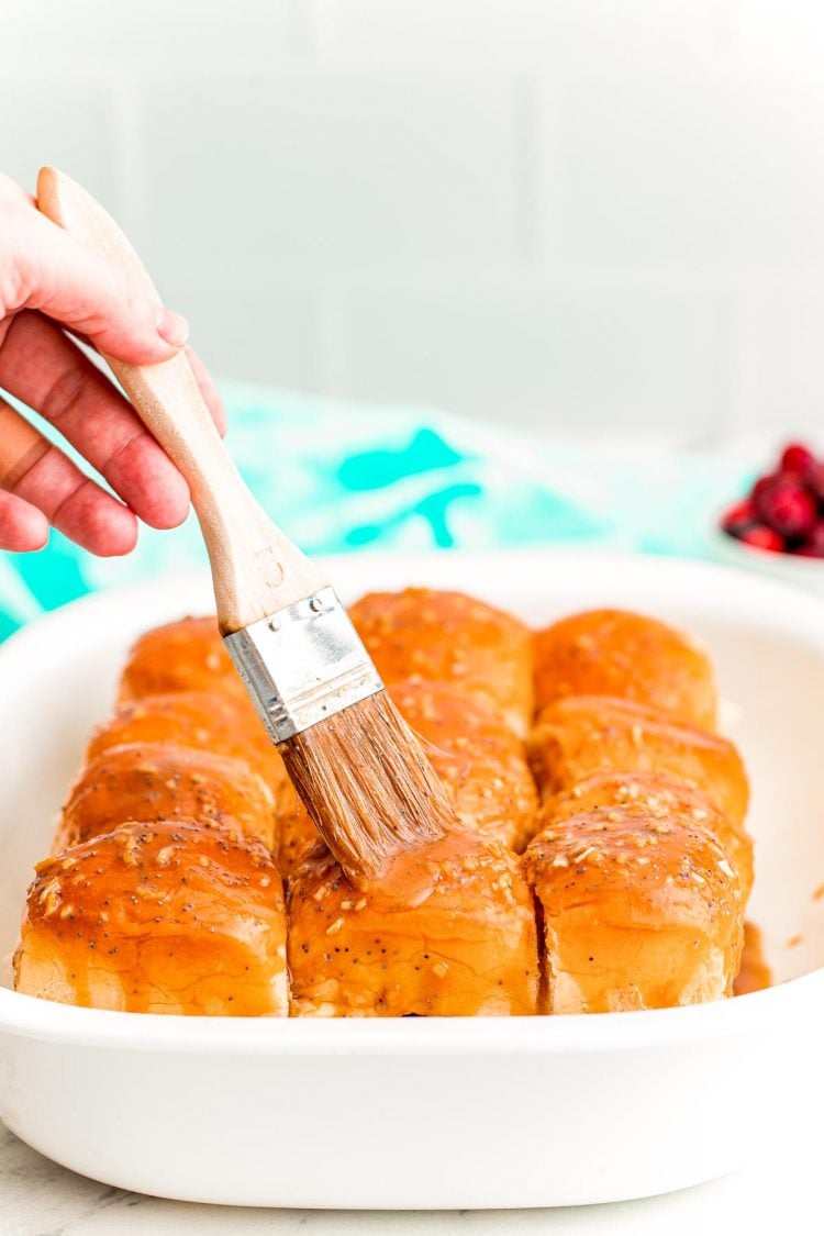 A woman's hand using a pastry brush to brush a butter mixture on top of the rolls of sliders in a white casserole dish.