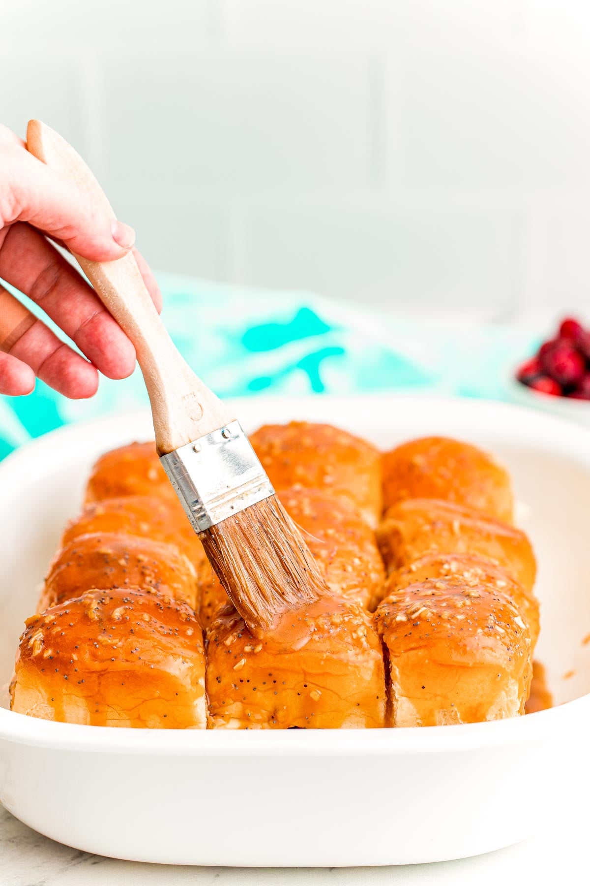 A woman's hand using a pastry brush to brush a butter mixture on top of the rolls of sliders in a white casserole dish.