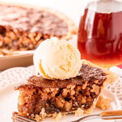 A slice of walnut pit on a white plate with a fork. Pitcher of maple syrup and the rest of the pie are in the background.