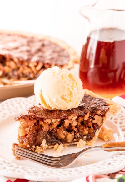 A slice of walnut pit on a white plate with a fork. Pitcher of maple syrup and the rest of the pie are in the background.