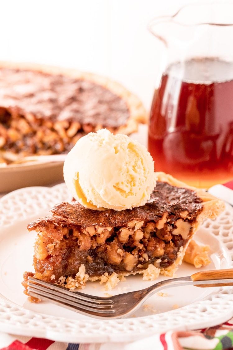 A slice of walnut pit on a white plate with a fork. Pitcher of maple syrup and the rest of the pie are in the background.