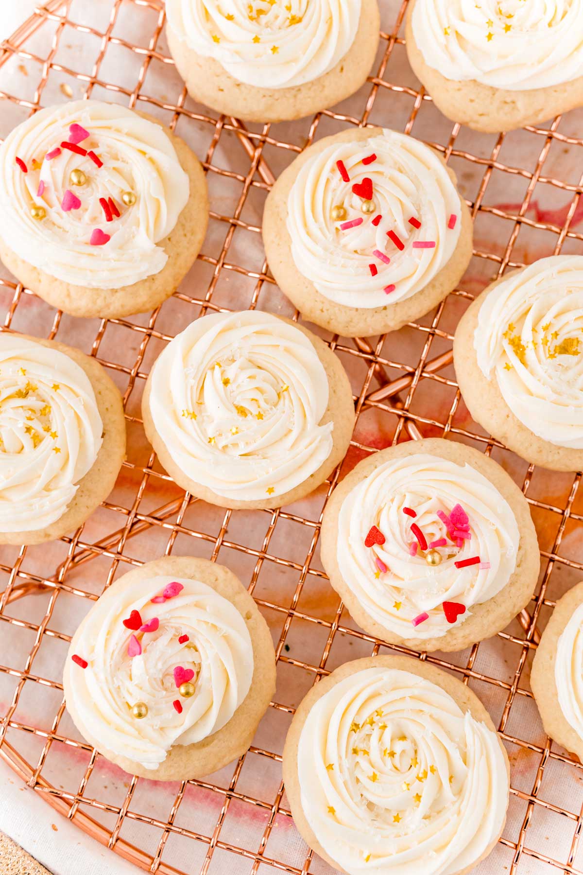 Overhead photo of sugar cookies with white frosting and sprinkles on a copper cooling rack.