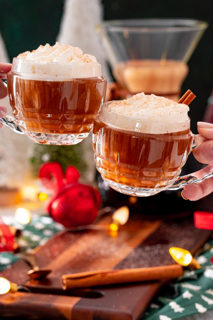 Two mugs of Christmas coffee being clinked over a table of holiday decorations.