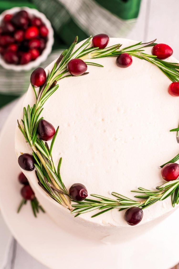 Top down photo of a cake with white frosting and topped with fresh cranberries and rosemary.