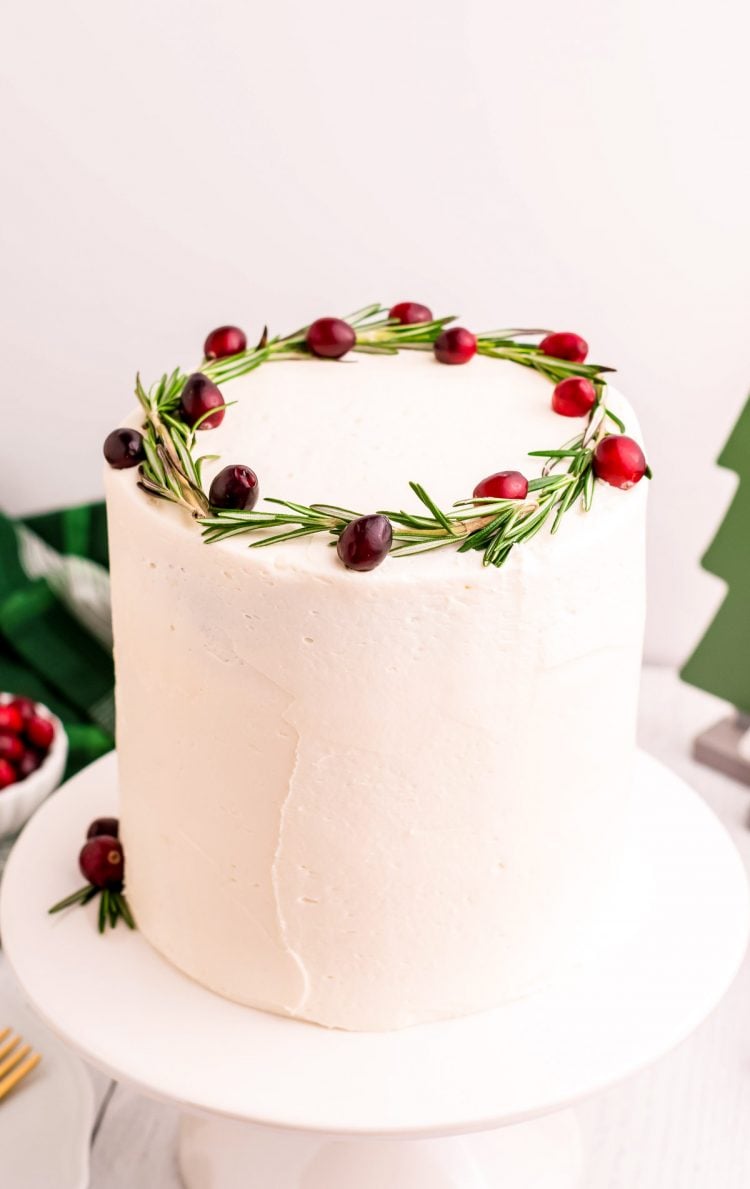 A White frosted layer cake topped with rosemary and cranberries on a white cake stand.