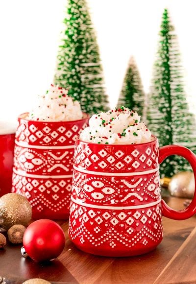 A red mug filled with eggnog hot chocolate and topped with whipped cream and christmas sprinkles.
