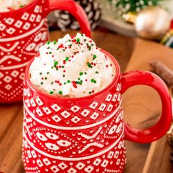 Close up photo of a red Christmas mug filled with hot chocolate and topped with whipped cream and sprinkles.