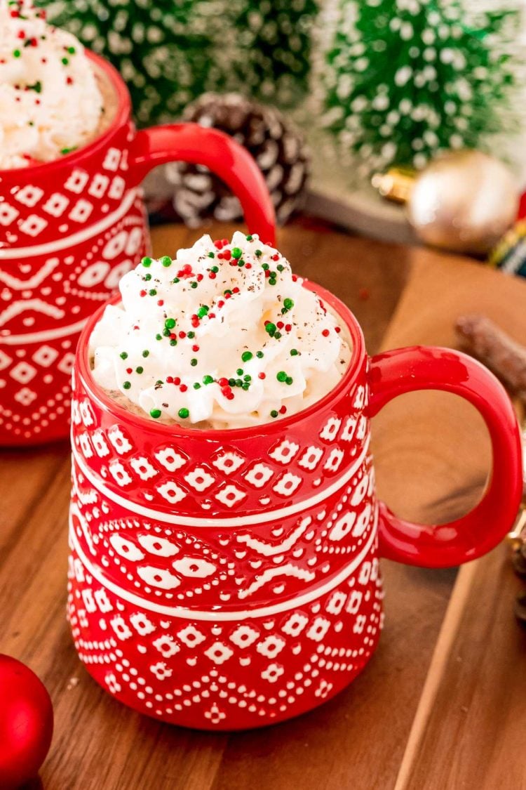 Close up photo of a red Christmas mug filled with hot chocolate and topped with whipped cream and sprinkles.
