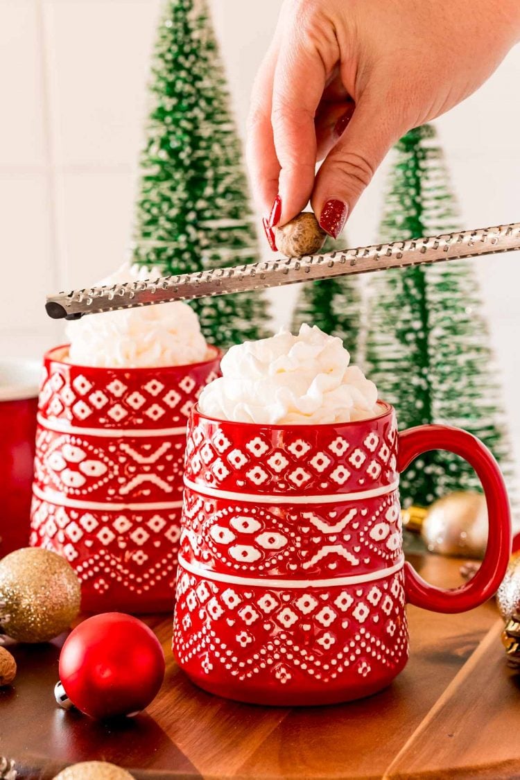 A woman's hand grating a nutmeg over hot chocolate in a red mug.