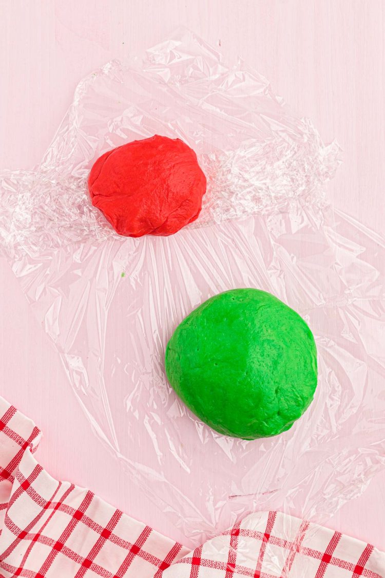 Red and green cookie dough being wrapped in plastic wrap on a pink table.