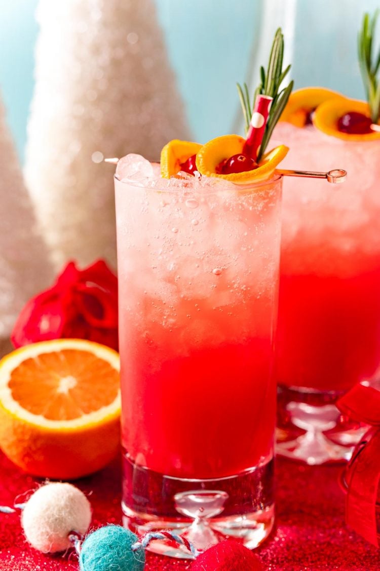 Close up photo of a tall glass filling with a red drink and garnished with oranges and cherries and rosemary.