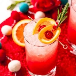 Close up photo of a tall glass filling with a red drink and garnished with oranges and cherries and rosemary.