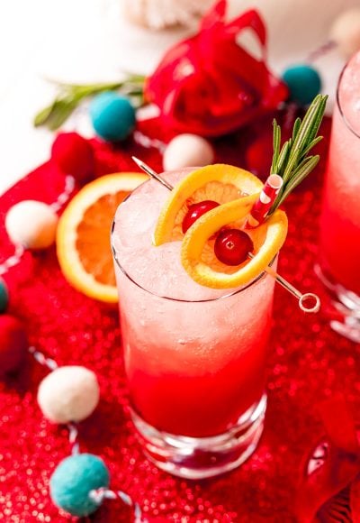 Close up photo of a tall glass filling with a red drink and garnished with oranges and cherries and rosemary.