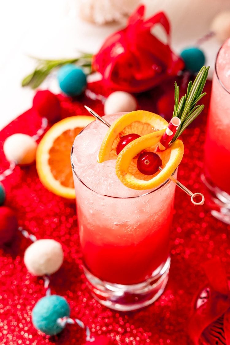 Close up photo of a tall glass filling with a red drink and garnished with oranges and cherries and rosemary.