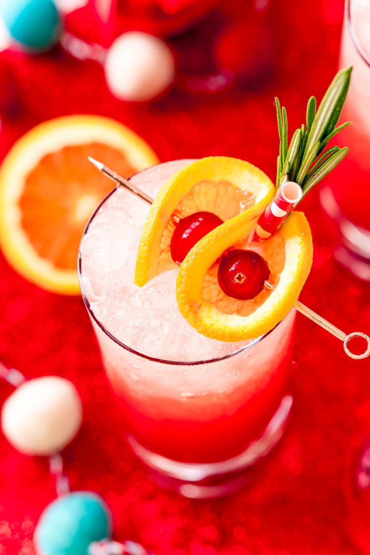 Close up photo of a tall glass filling with a red drink and garnished with oranges and cherries and rosemary.