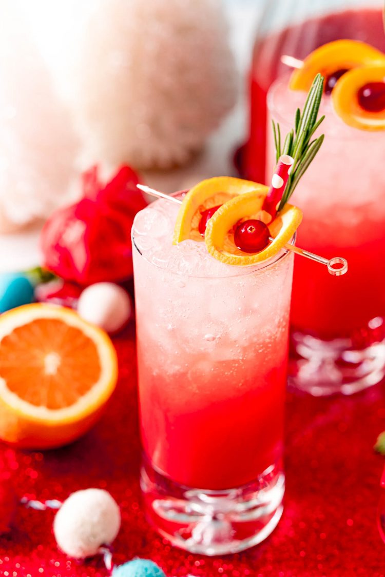Close up photo of a tall glass filling with a red drink and garnished with oranges and cherries and rosemary.