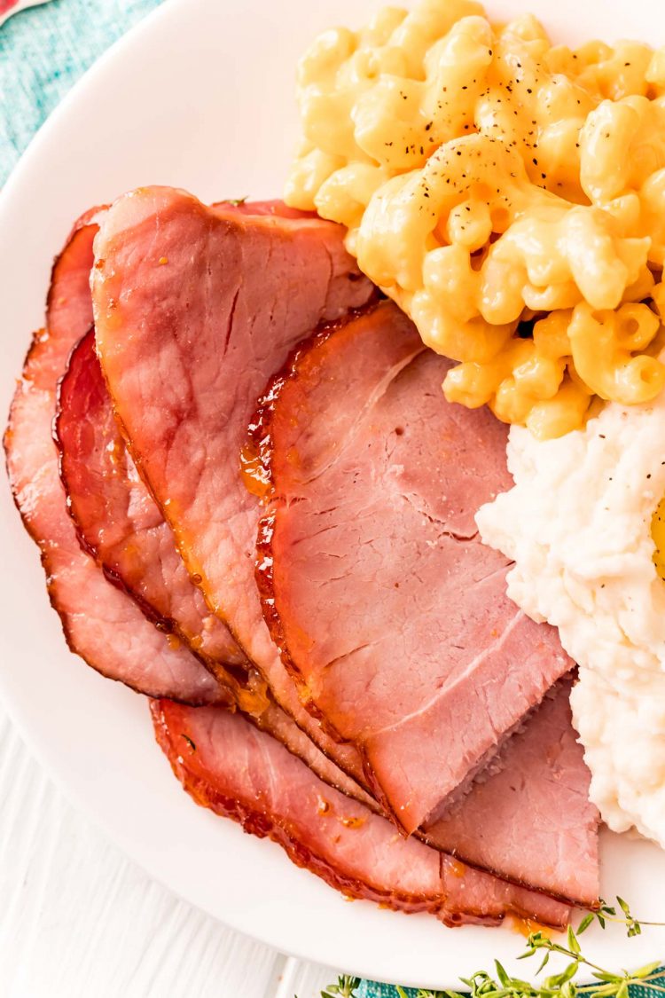 Close up photo of slices of ham on a plate with mac and cheese and mashed potatoes.