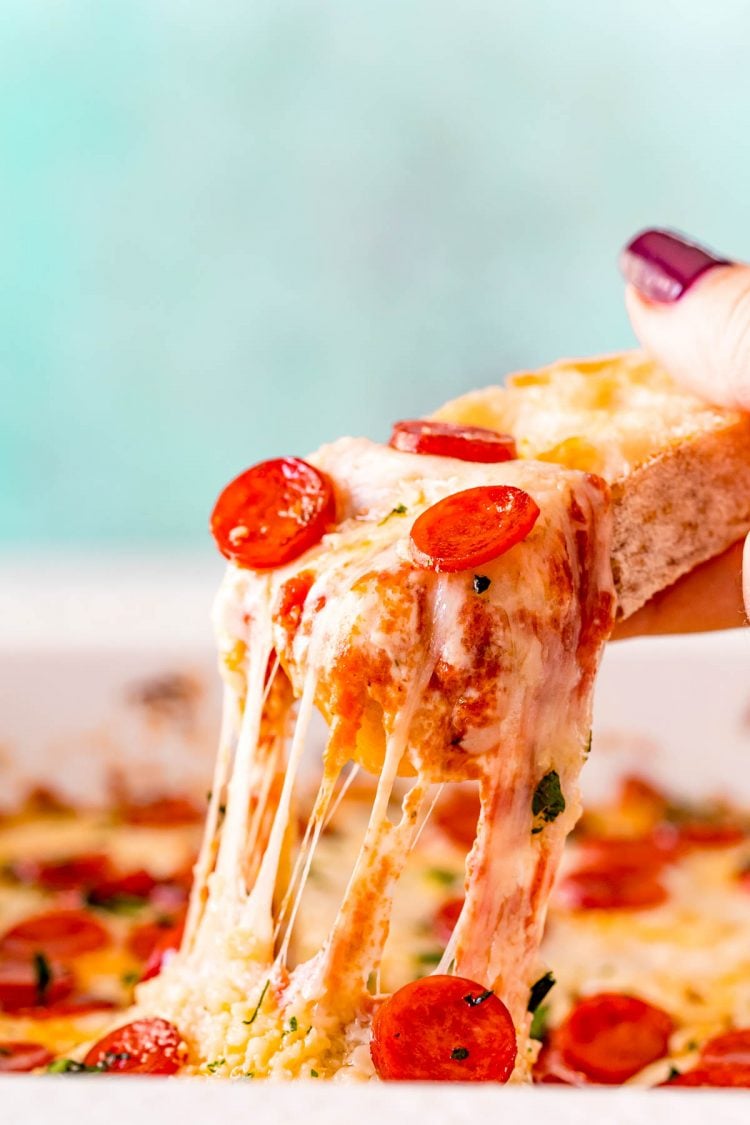 A woman's hand scooping pizza dip our of a white baking dish with a piece of bread.